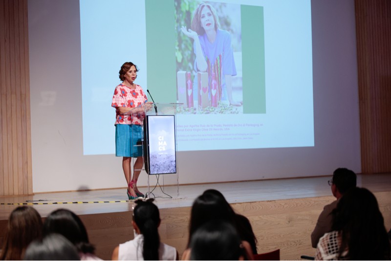 Speaker presents on stage with audience, large screen projection in background.